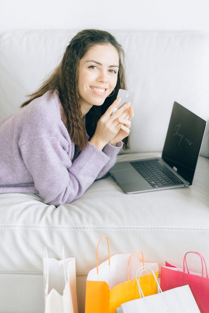 Smiling woman using her credit card for online shopping while lounging on a sofa with colorful shopping bags around.